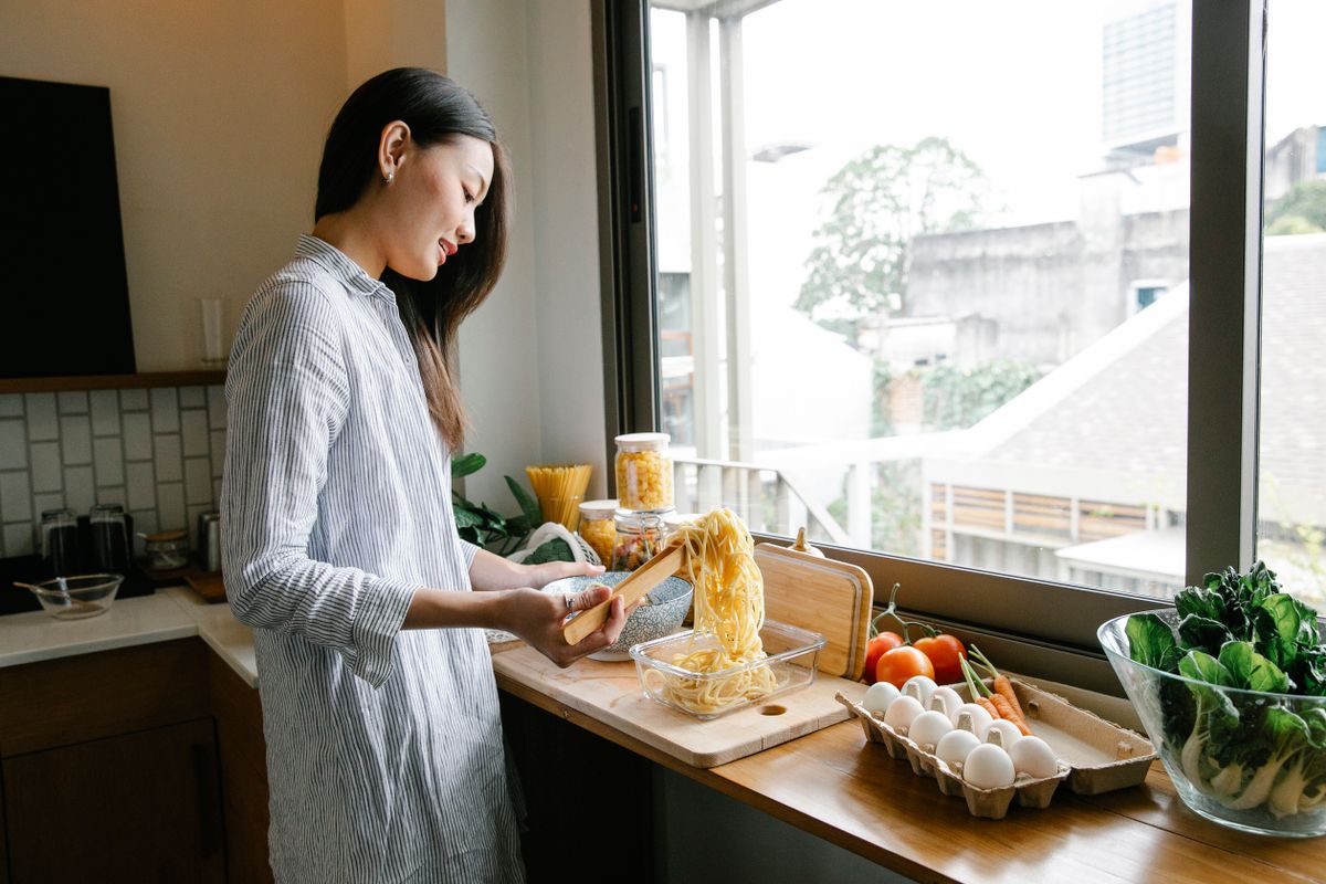 Side view of Asian female cook putting long spaghetti into glass container while standing at counter with various ingredients in kitchen