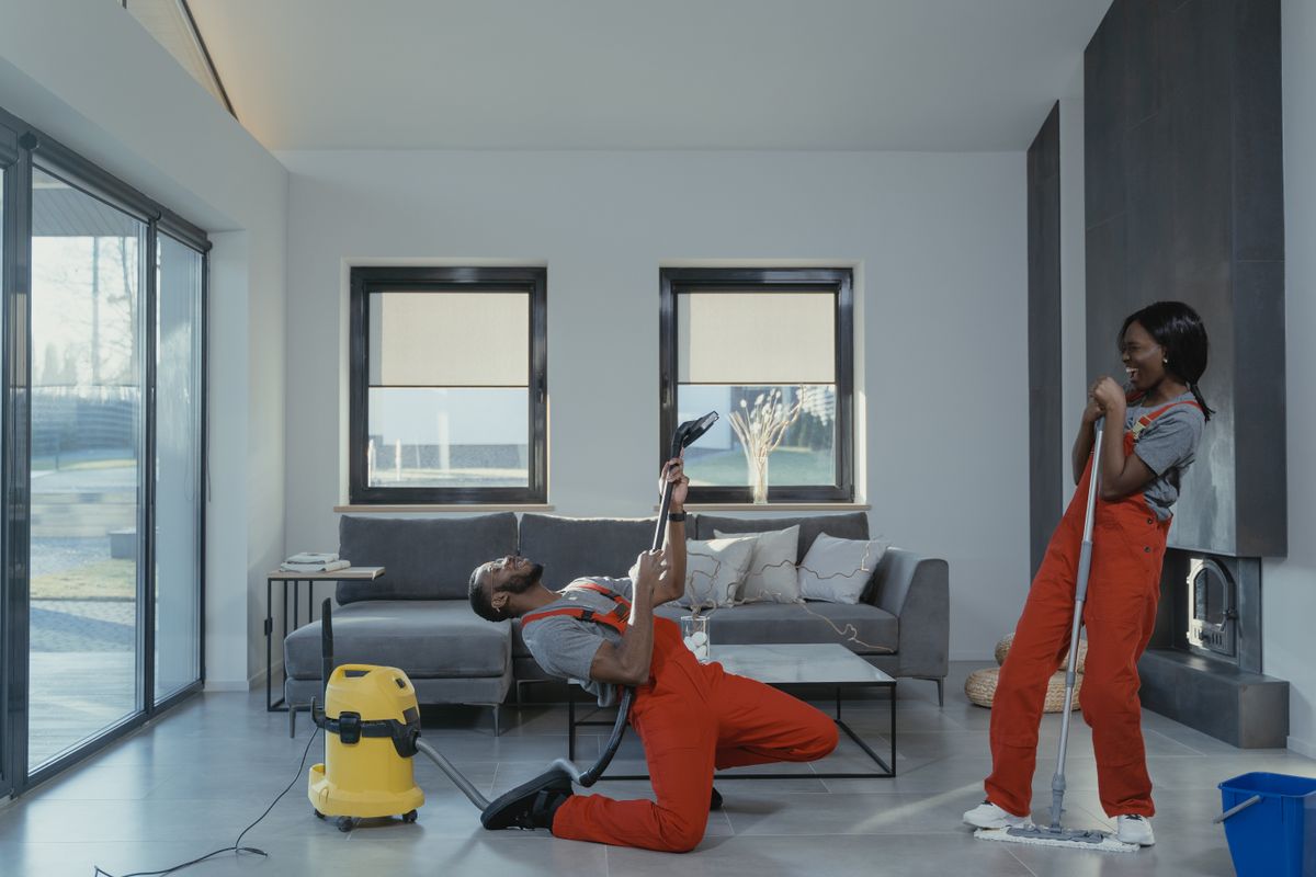 Fun and engaging cleaning session with a man and woman in overalls using cleaning tools in a modern living room.