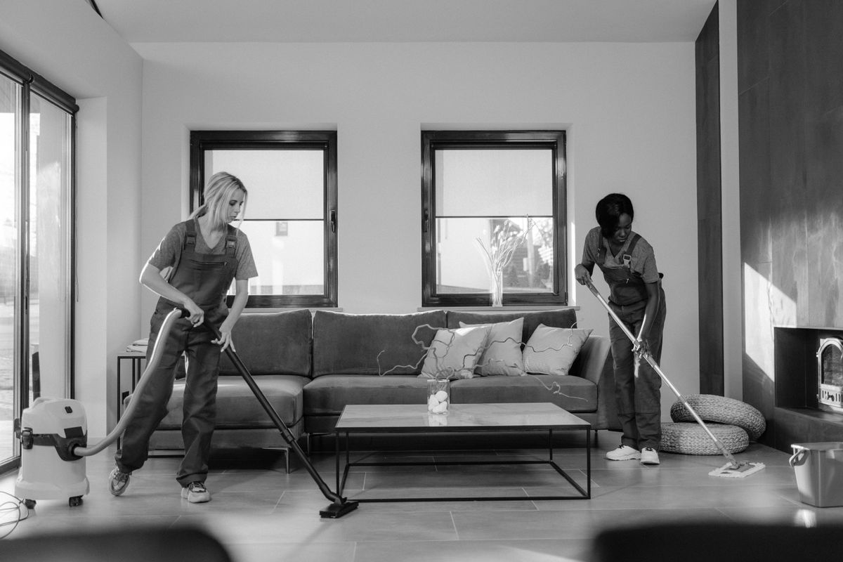 Two women in work clothes cleaning a modern living room using a vacuum and mop.
