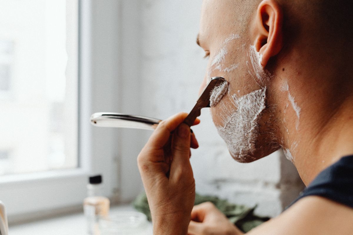 Young man using a straight razor to shave his face in the bathroom.