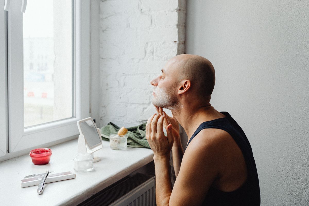 A bald man applying shaving cream to his chin while looking out the window
