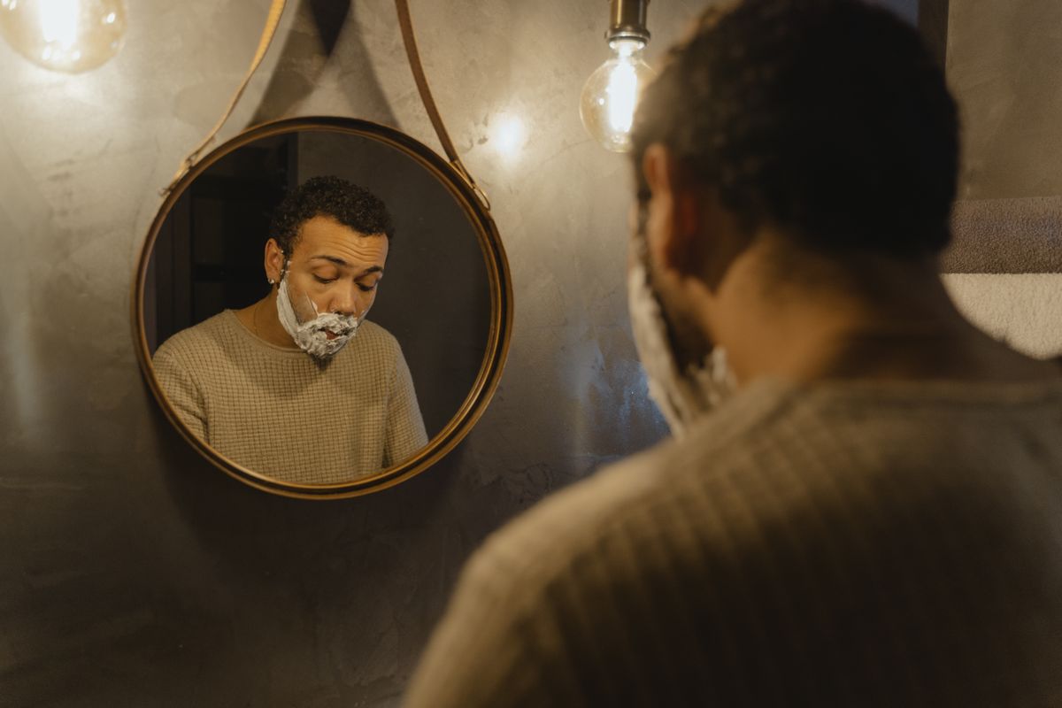 Man applying shaving cream while looking in round mirror in bathroom.