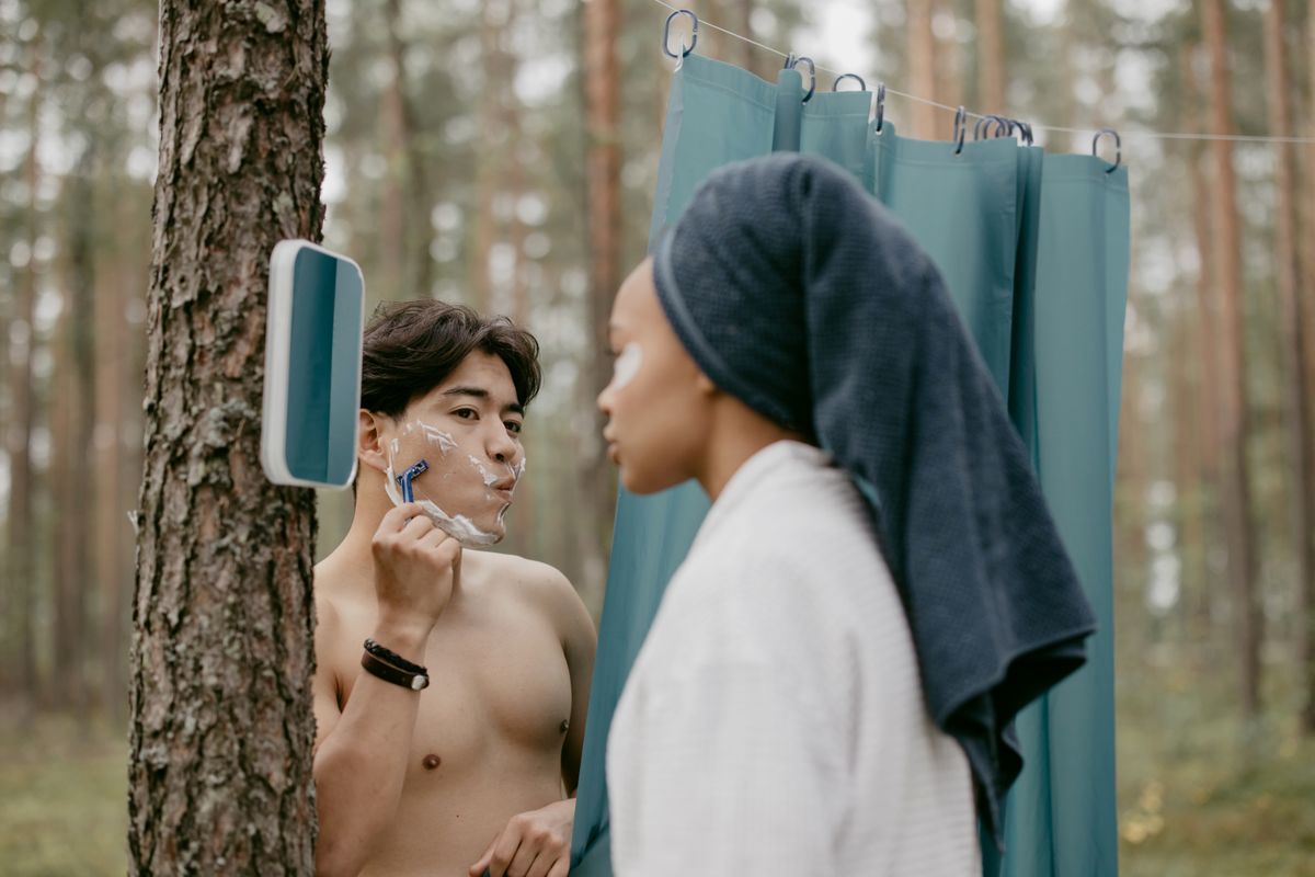 A man shaving outdoors using a tree-mounted mirror as a woman applies face cream next to him.