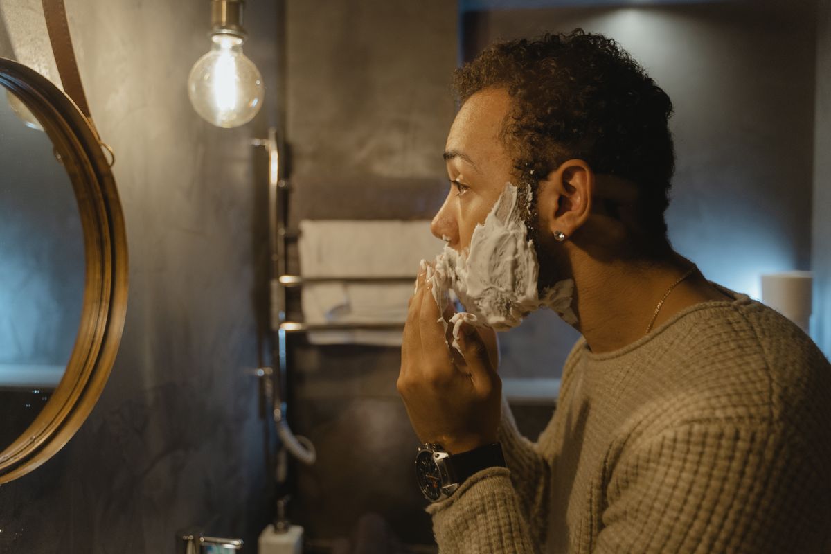 Side view of a man applying shaving cream while looking in the bathroom mirror.