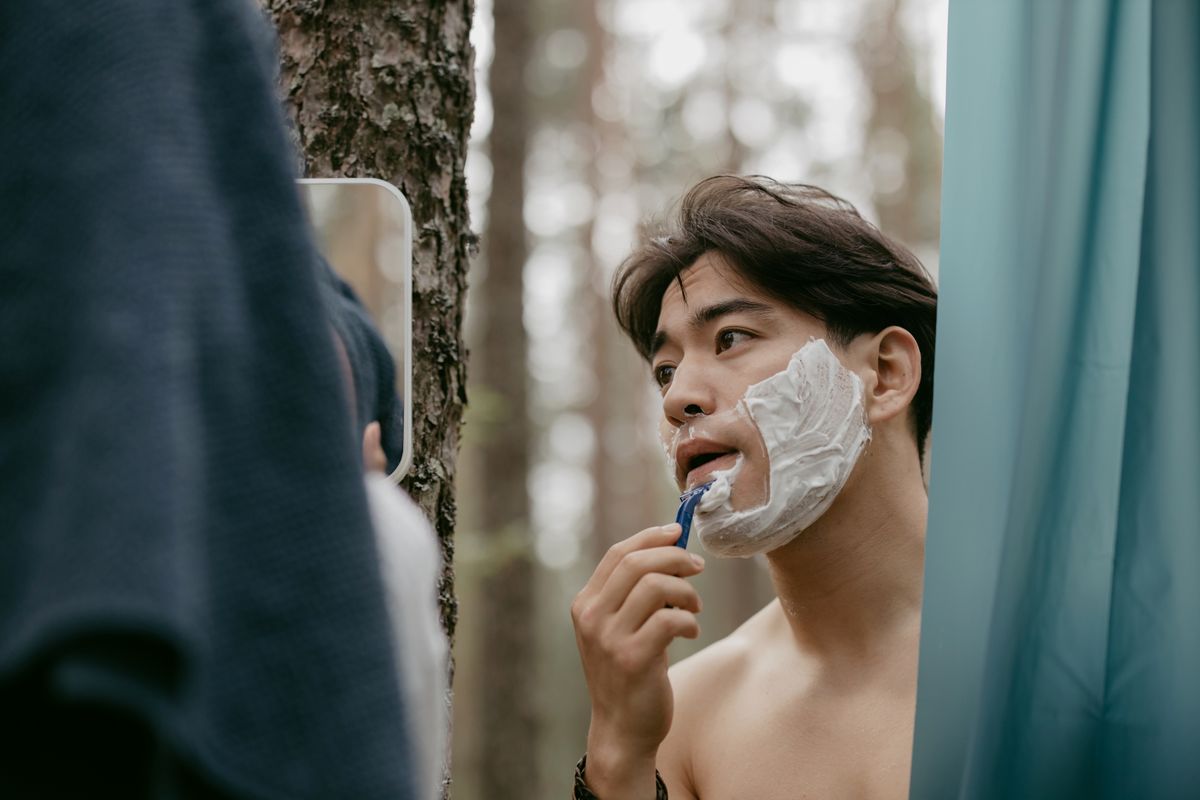 Asian man shaves his face outdoors in a forest, using mirror and razor.
