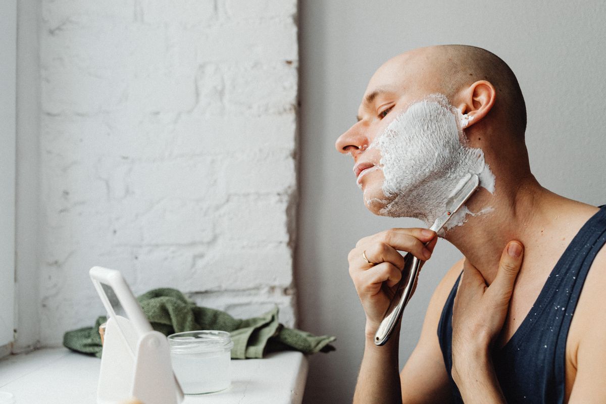 Bald man applying shaving cream and using razor in bright indoor setting.