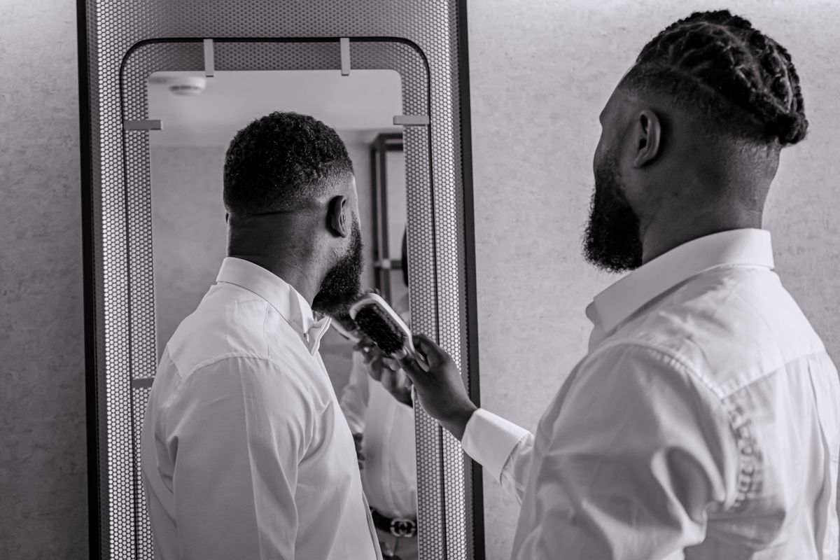 A man in a white shirt brushes his beard while standing in front of a mirror indoors.