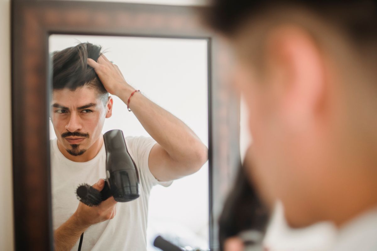 Adult man styles his hair using a hairdryer while looking at his reflection, capturing a modern grooming routine.