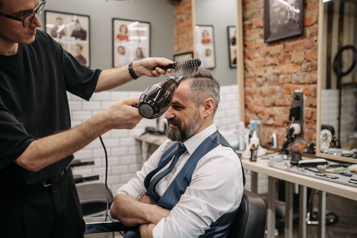 An elderly man gets his hair blow-dried by a barber in a modern barbershop setting.