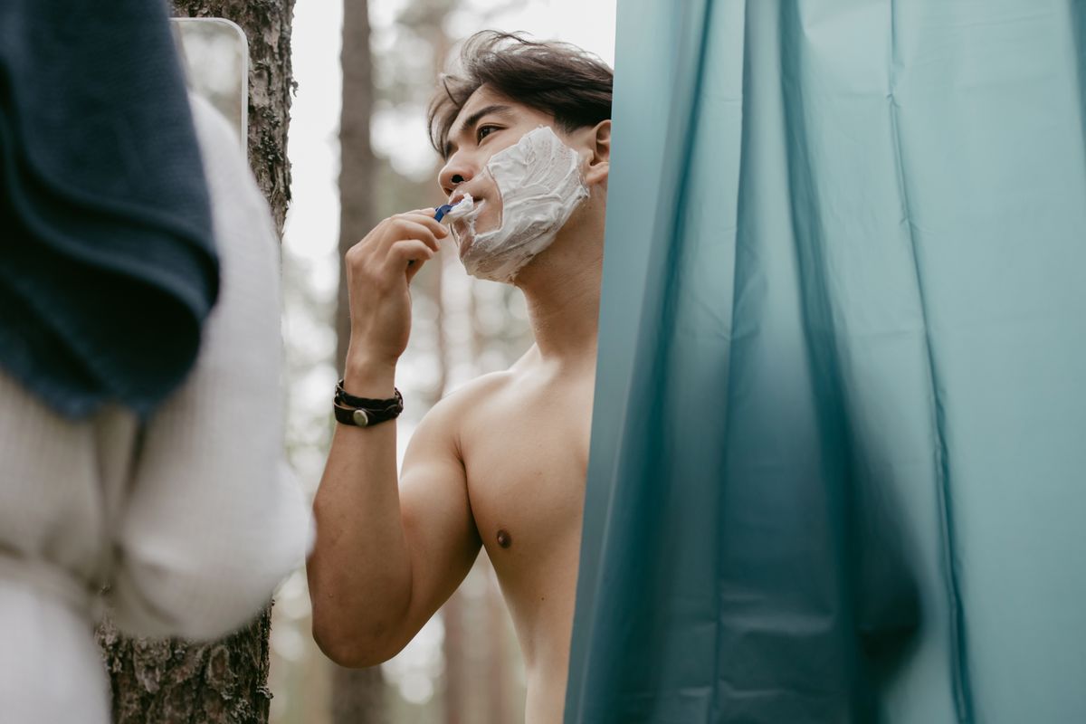 Asian man shaving with foam, using a razor outdoors near a shower curtain.