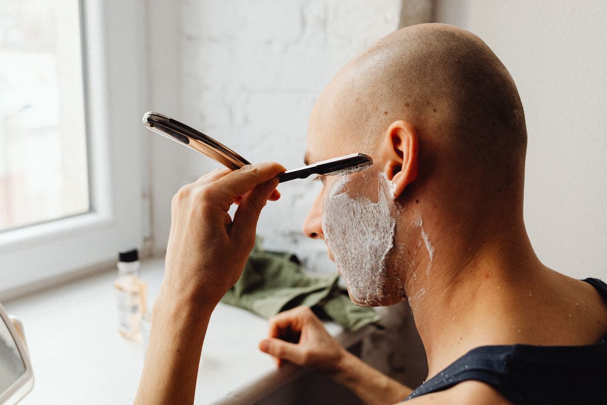 Adult man shaving with straight razor and shaving cream in a light-filled room.