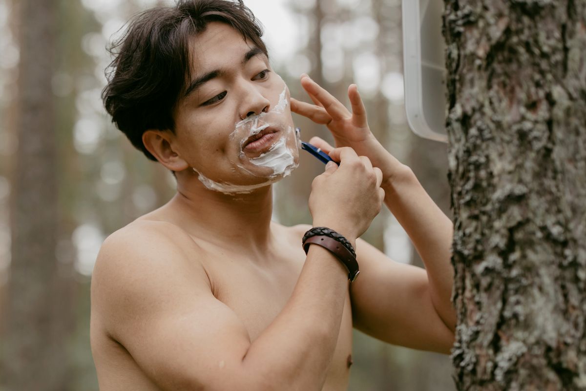Shirtless Asian man shaving with razor in forest, embracing a natural grooming routine.