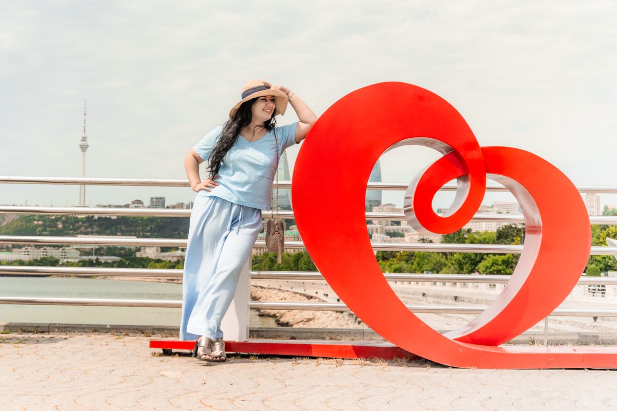 Woman in blue outfit posing with a heart sculpture in Baku, Azerbaijan. Daytime shot with a scenic backdrop.