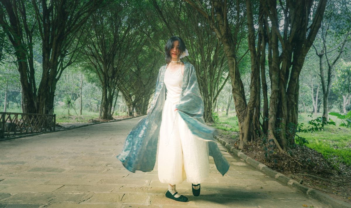 A serene scene of a woman in a flowing dress walking along a forest pathway under an arch of trees.