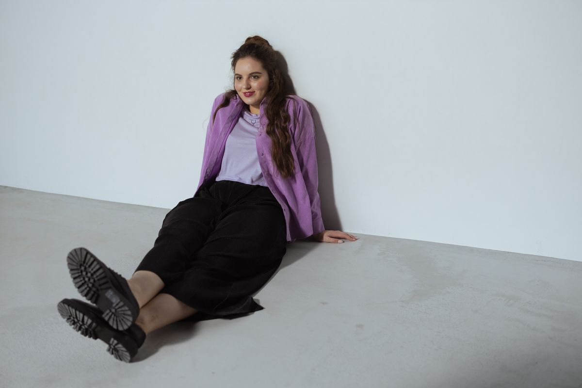 Young woman wearing a stylish purple top and black pants, sitting and smiling in a modern studio setup.