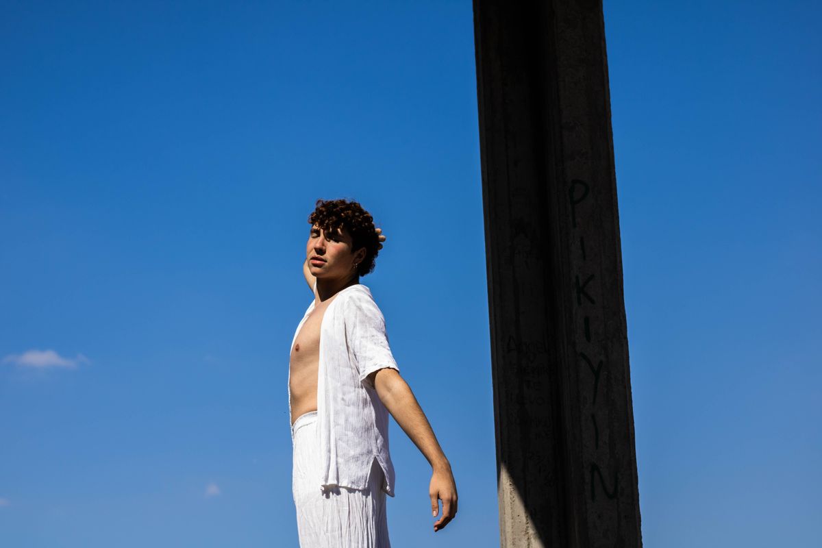 A model poses shirtless outdoors under a vibrant blue sky, showcasing fashion and confidence.
