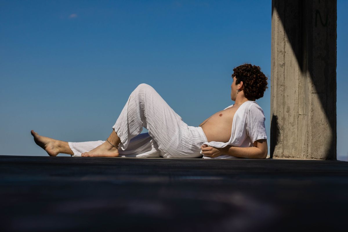 A man in white clothes relaxes barefoot against a column under a clear blue sky.