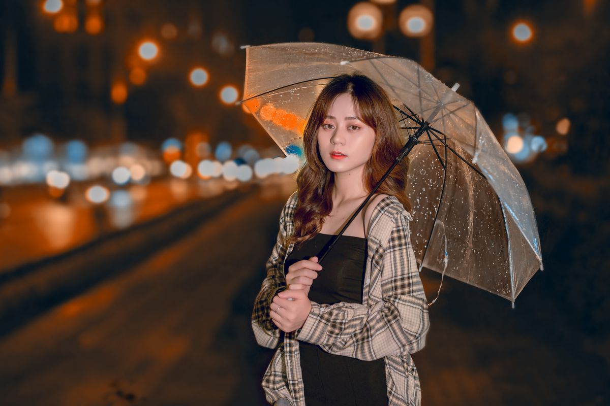 Asian woman holding a transparent umbrella amidst bokeh lights on a rainy night street.