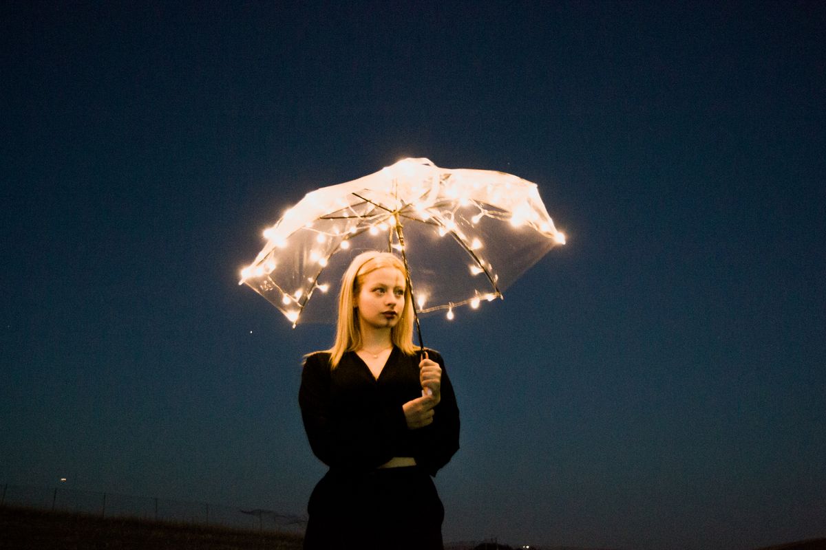 A young woman stands at night holding an umbrella decorated with string lights, creating a dreamy effect.