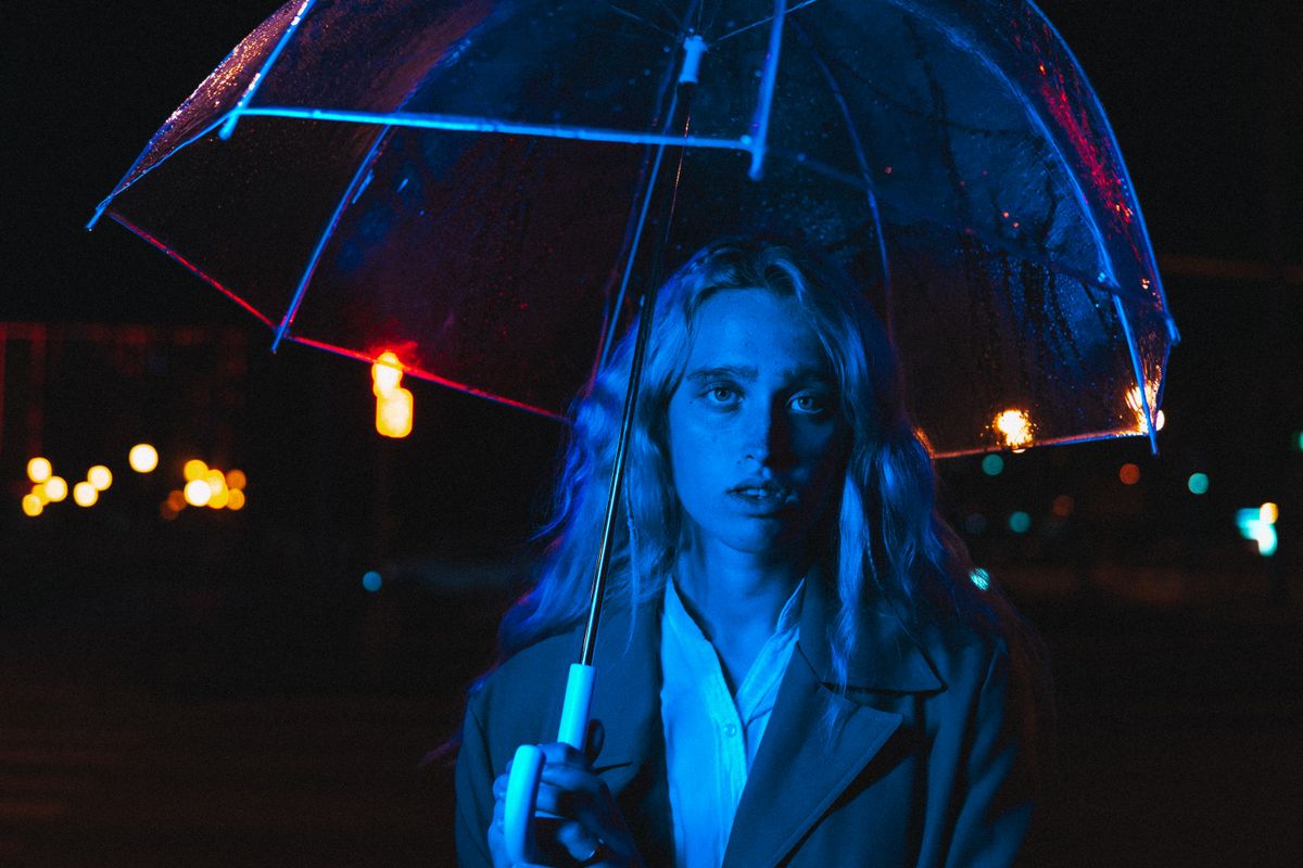 Dramatic portrait of a woman under a glowing umbrella, bathed in blue and red night lights.