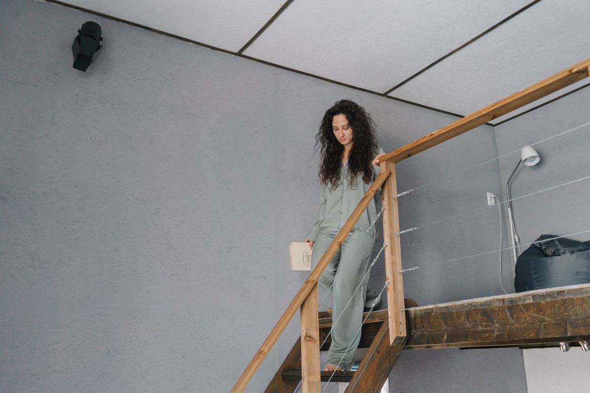 Woman with curly hair wearing pajamas descends wooden stairs indoors holding a book.