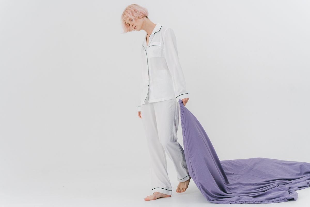 A woman wearing pajamas drags a purple blanket in a minimalist studio setting.