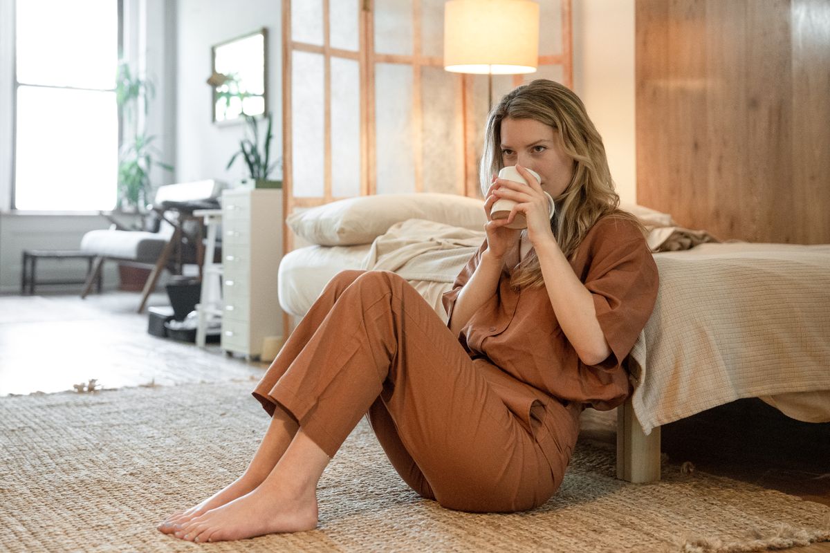 Woman in casual outfit relaxing on bedroom floor with coffee mug, enjoying a tranquil morning.