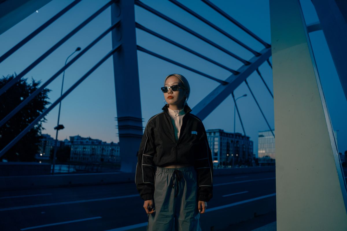Asian woman wearing a black jacket stands on a bridge at twilight, exuding a cool and modern vibe.