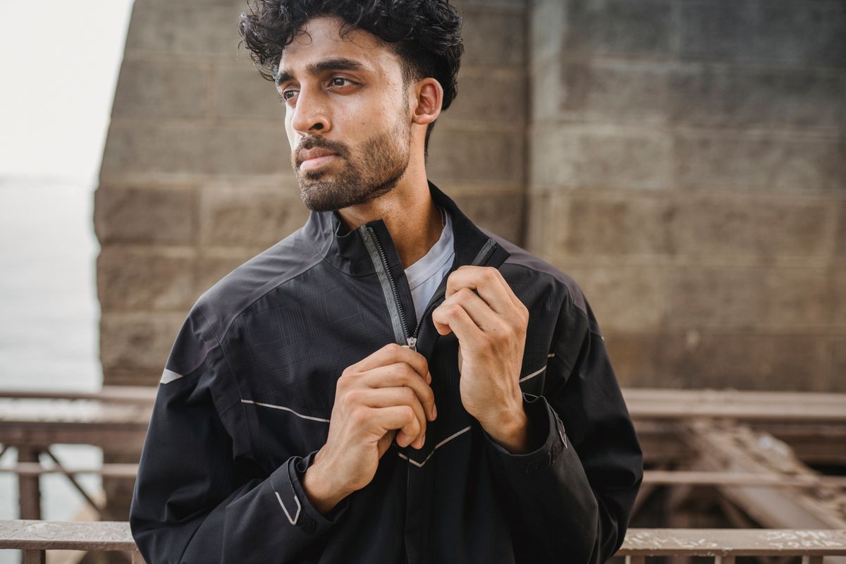Stylish young man zipping up jacket beneath an urban bridge, exuding confidence.