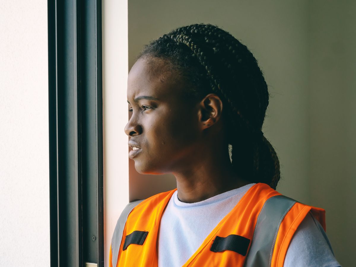 Profile of an adult African American woman wearing a reflective orange vest, looking out a window.