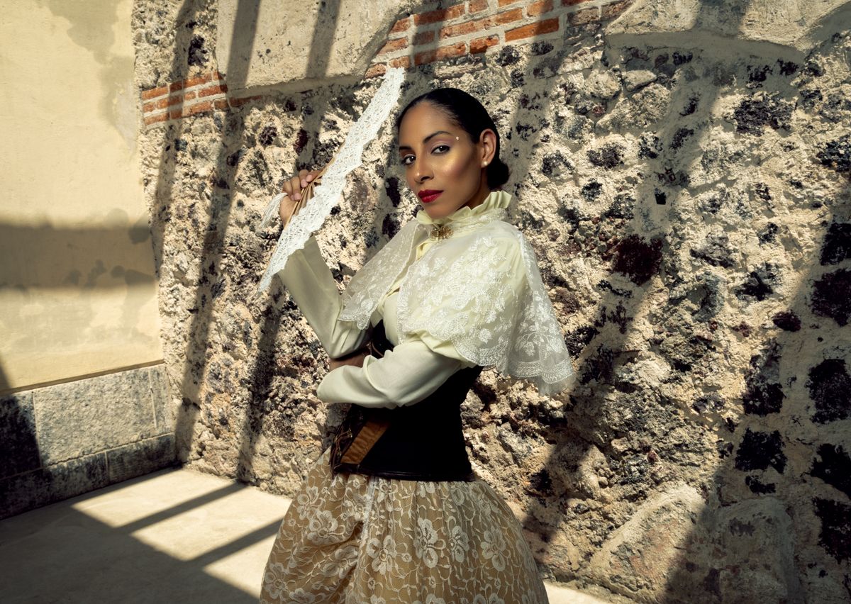 A young woman poses elegantly in a vintage dress, holding a lace fan against a textured stone wall.