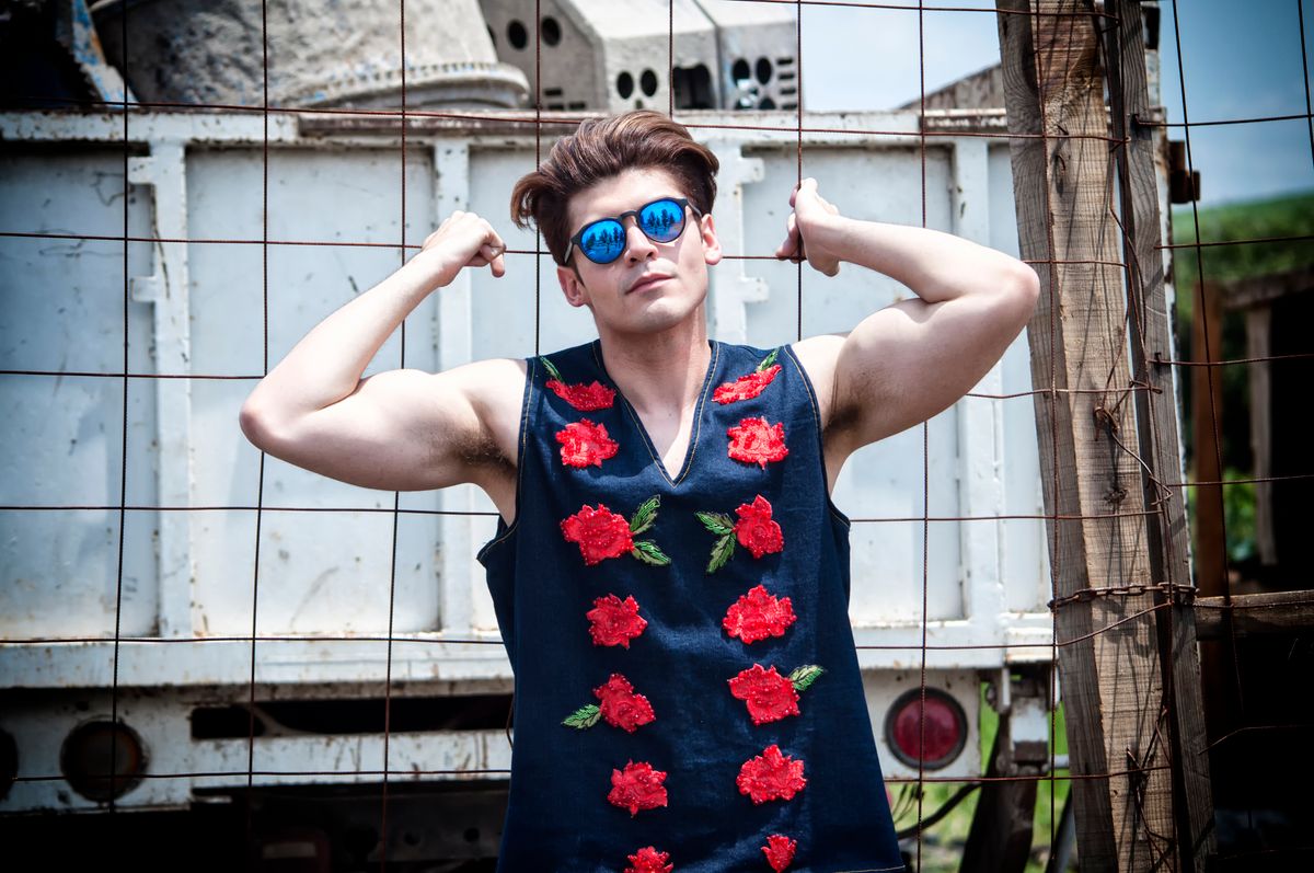 Confident man wearing a floral embroidered vest and sunglasses, posing outdoors against a rustic backdrop.