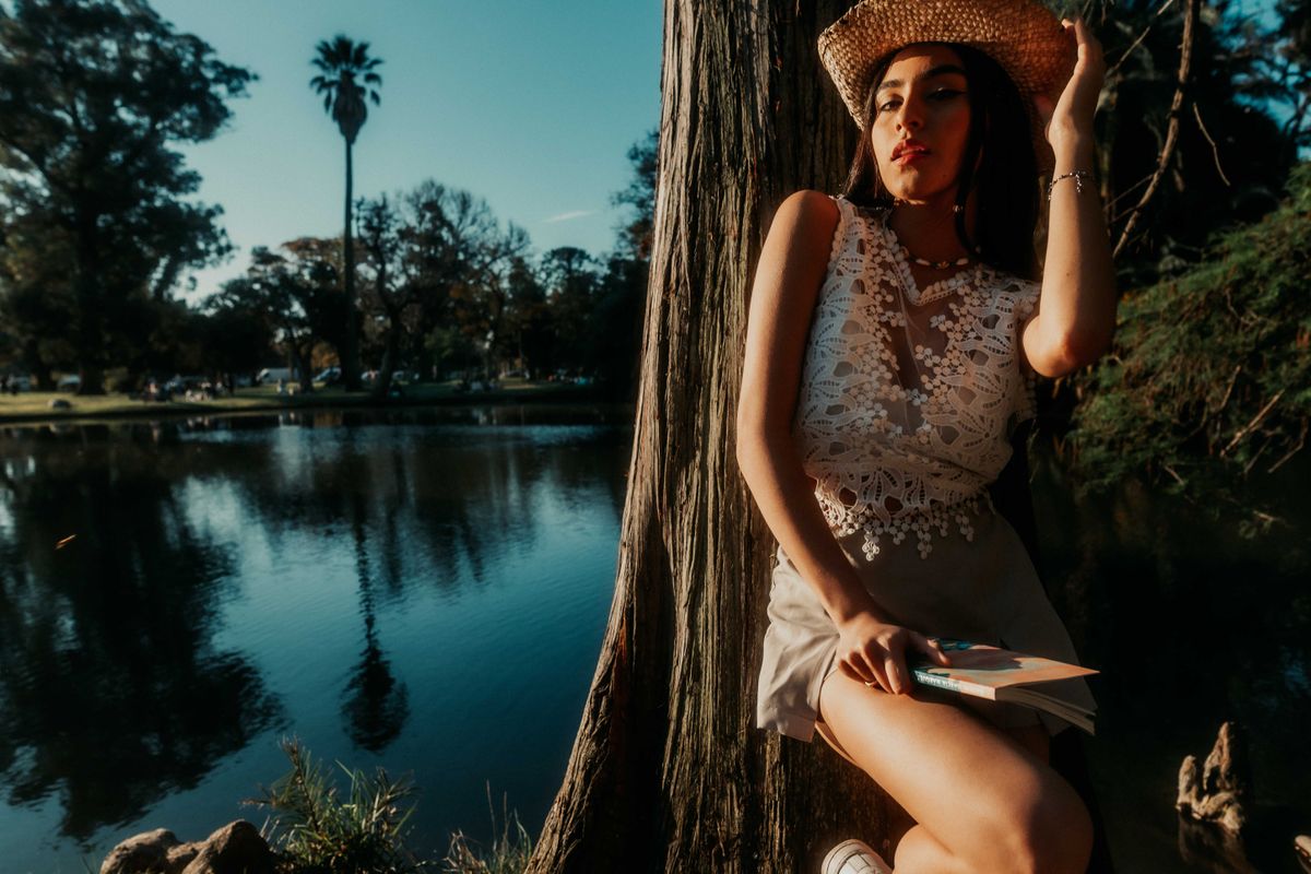 A woman in a lace dress and hat, holding a book, relaxes by a scenic lakeside in warm evening light.