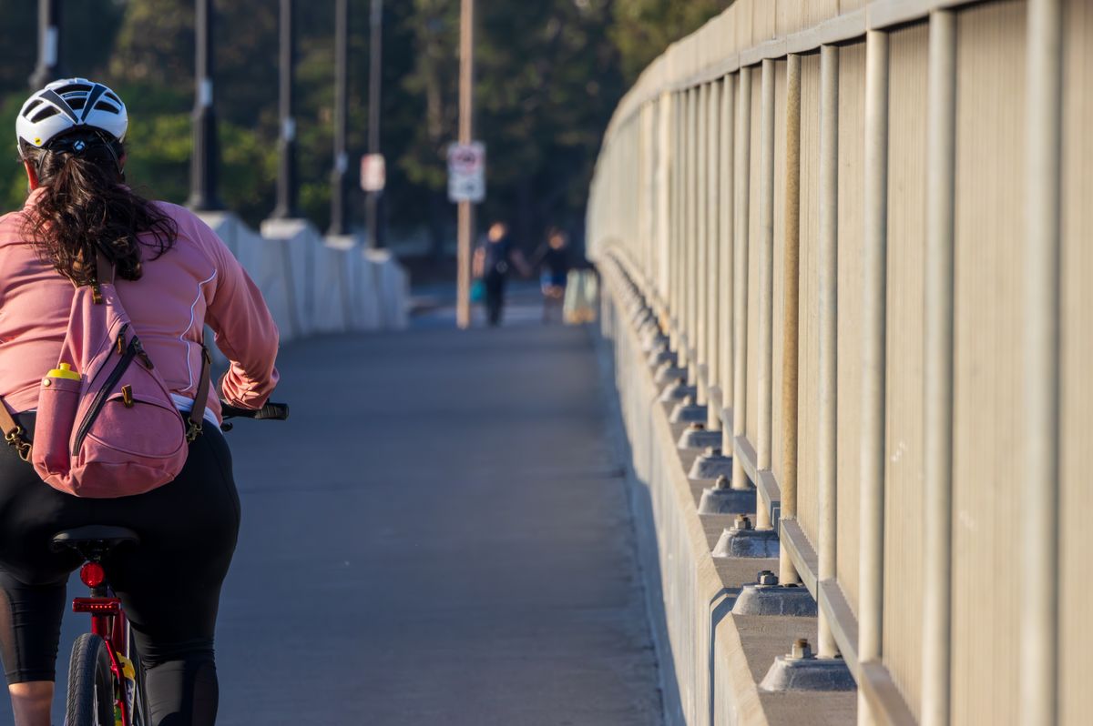 A woman cycling with a backpack along a city bridge during the day, wearing a helmet.