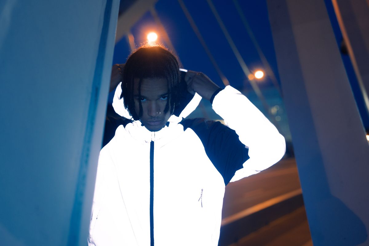 A man wearing a reflective white hoodie stands under bridge lights at night, creating an urban vibe.