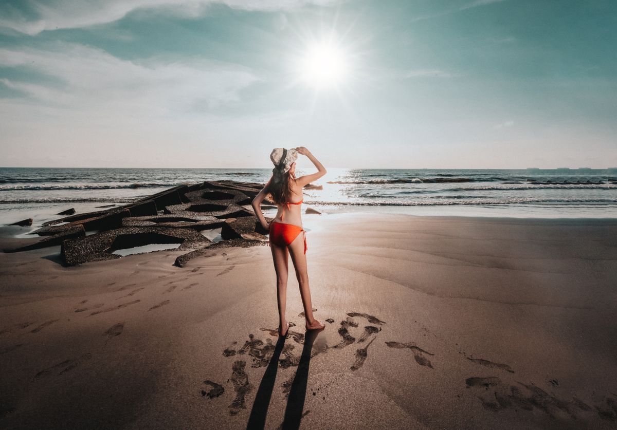 A woman in a red bikini stands on a sandy beach looking at the ocean under bright sunlight.