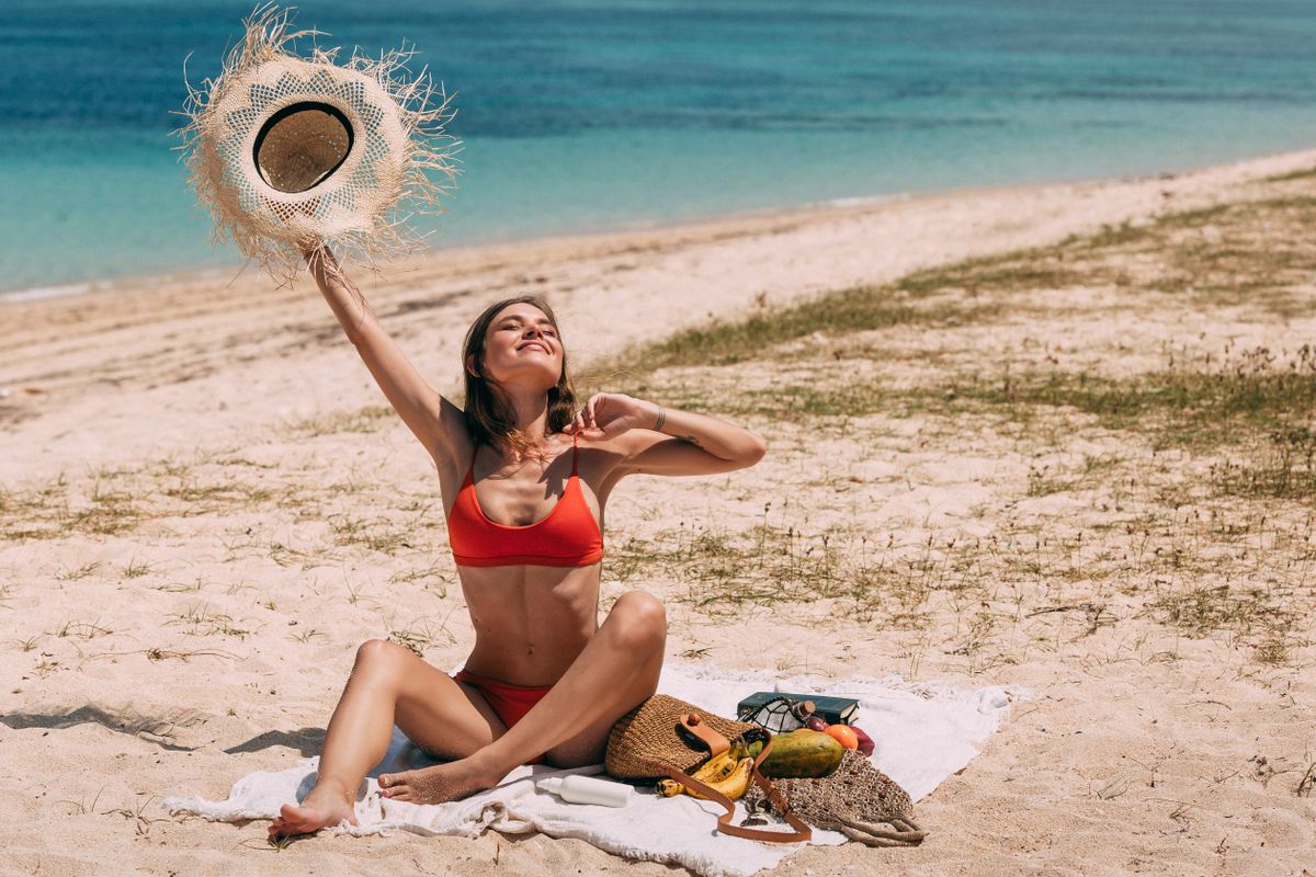 Woman in red bikini enjoys a sunny day on a sandy beach, holding a straw hat.