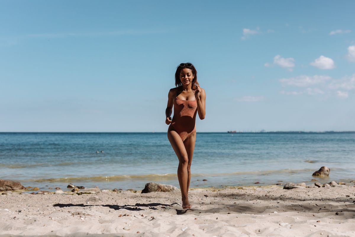 A woman in a swimsuit runs joyfully along a sandy beach under a clear blue sky.
