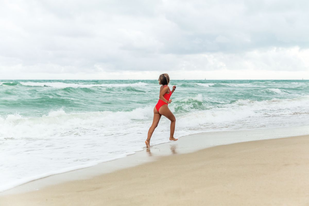 Full body back view of young tanned slim female traveler in red one piece body suit running on wet sand on coast of wavy sea under overcast sky covered with clouds