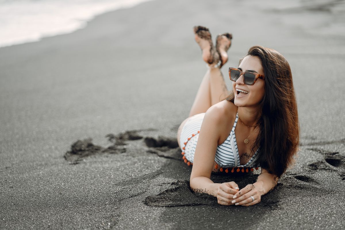 Full length cheerful barefoot female in swimwear and sunglasses lying on stomach on wet sandy beach near waving sea