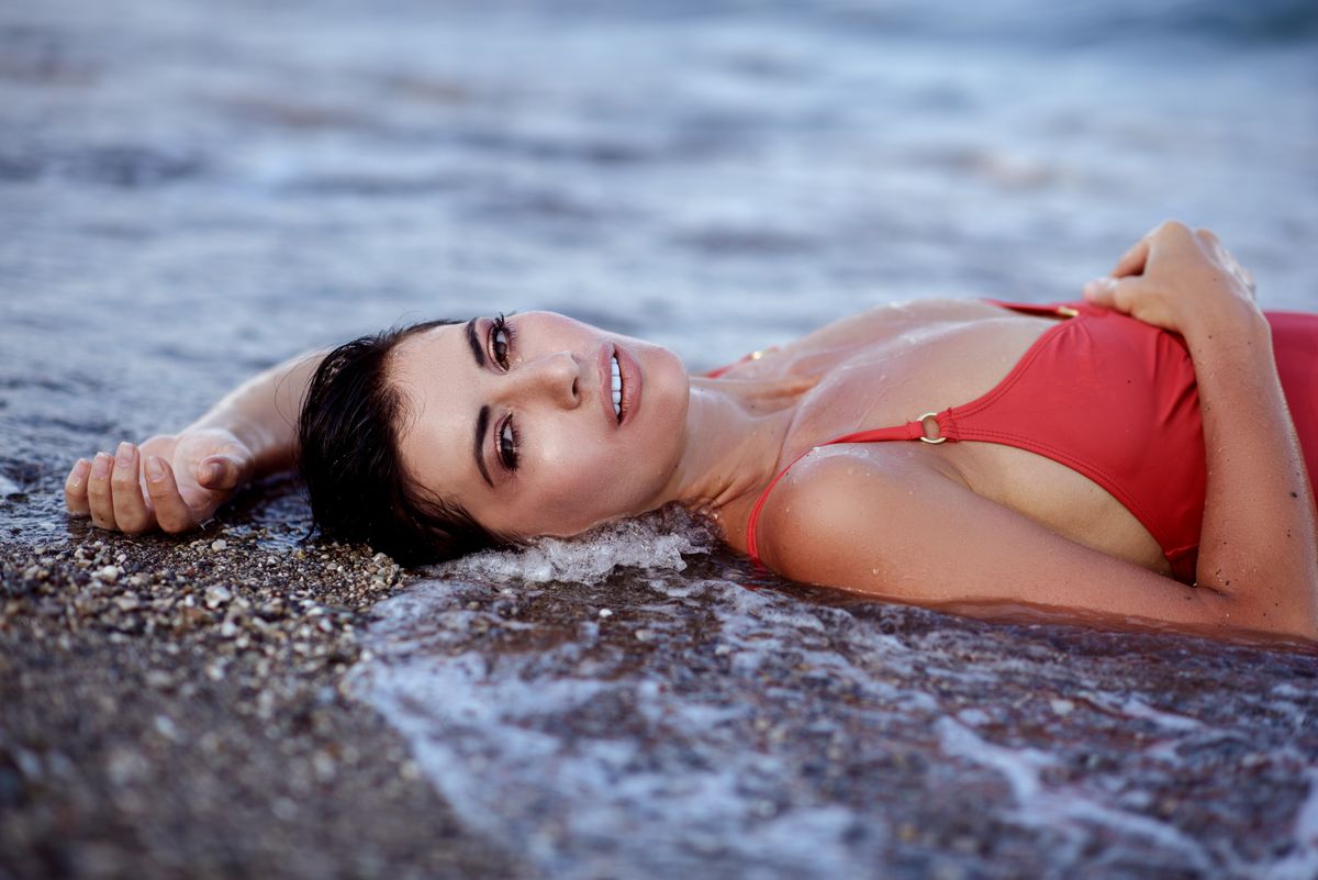 Sensual female with dark hair in red swimsuit lying on beach in water of ocean and looking at camera in daylight