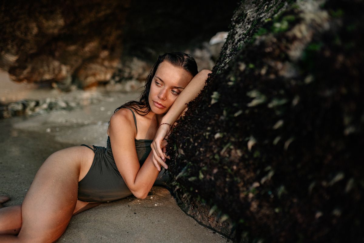 Young female in swimwear lying on sandy beach in sea water while leaning on slope of mountain relaxing after swimming