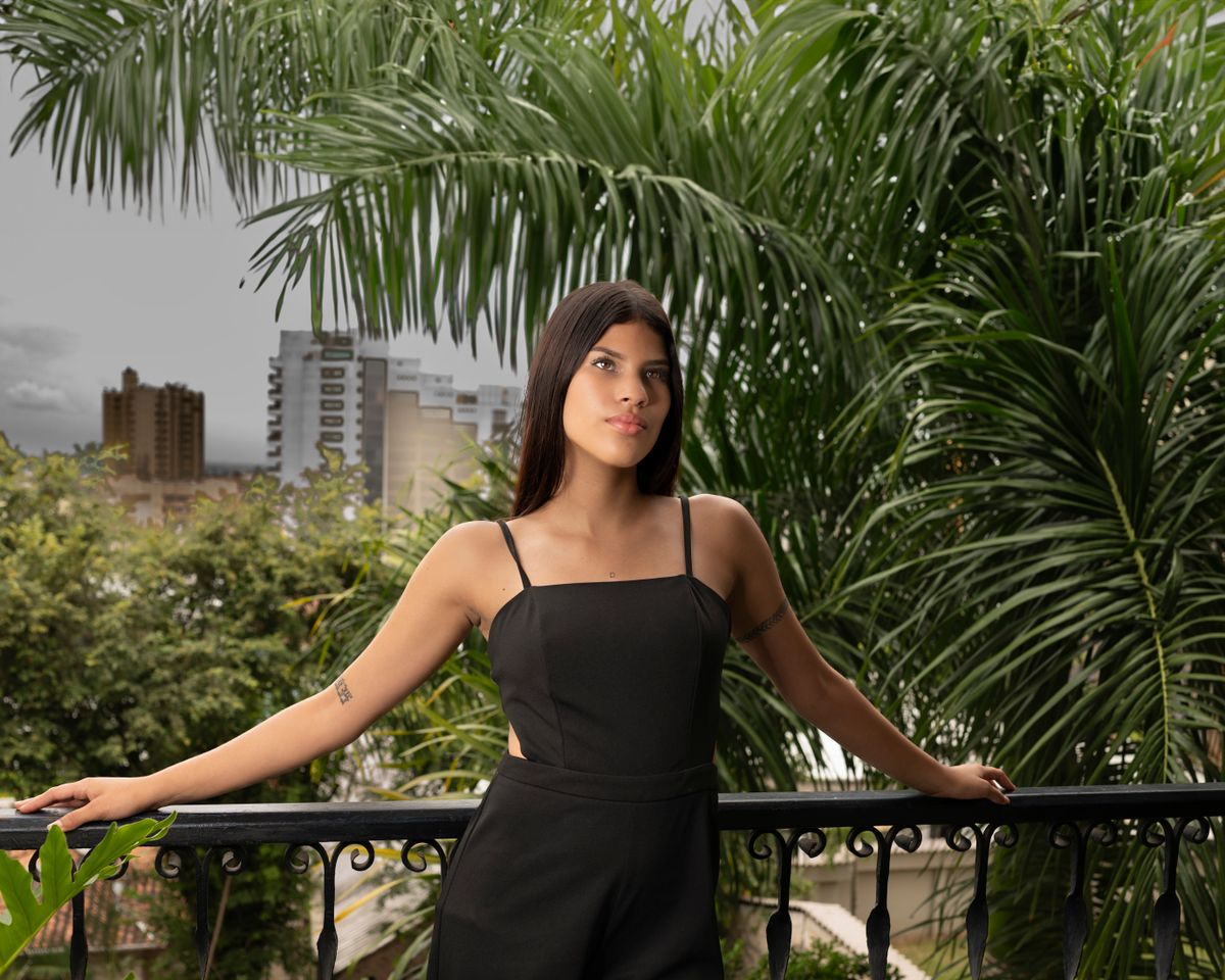 Chic brunette woman in black jumpsuit posing on a balcony with urban palm backdrop.