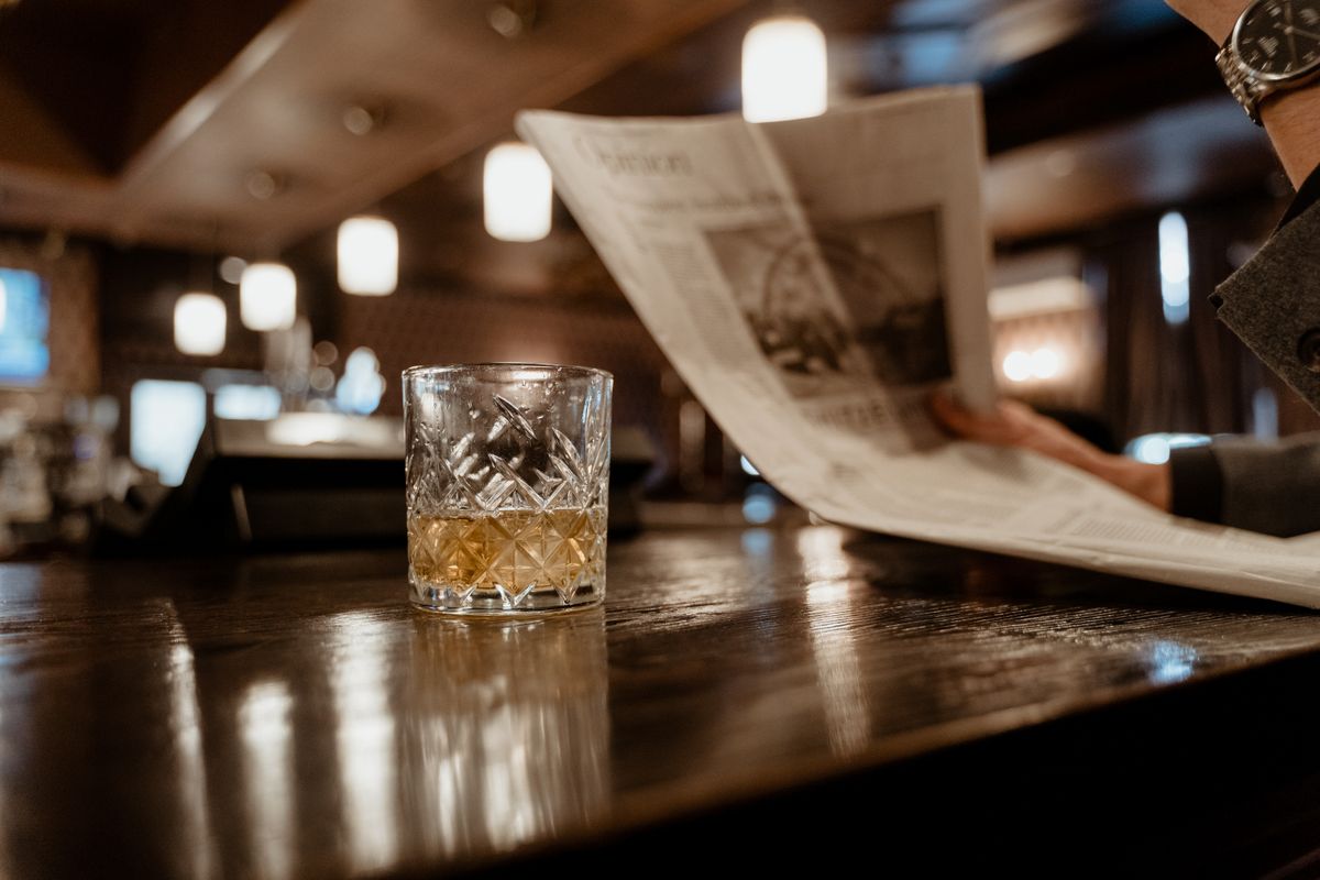 A close-up of a whiskey glass on a bar counter with a person reading a newspaper in the background.