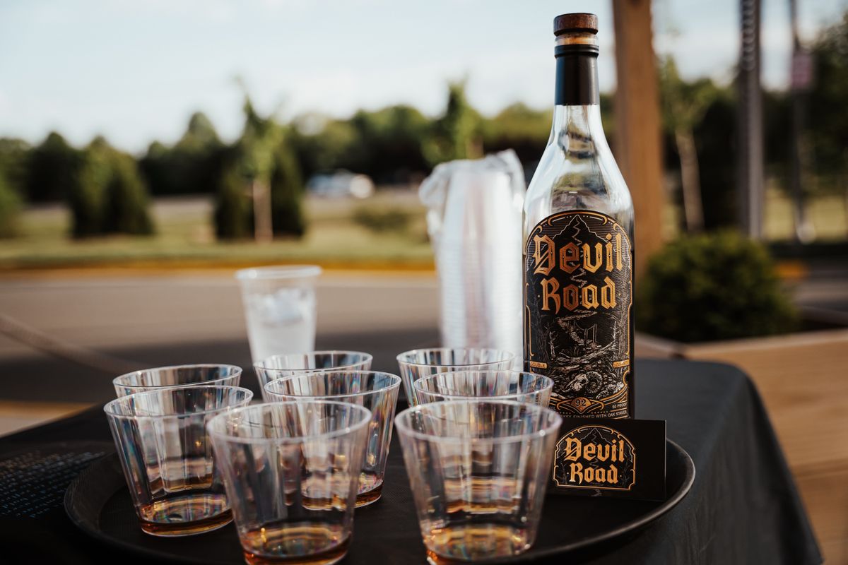 Whiskey bottle labeled 'Devil Road' with glasses set on a table outdoors. Perfect for a summer party setting.