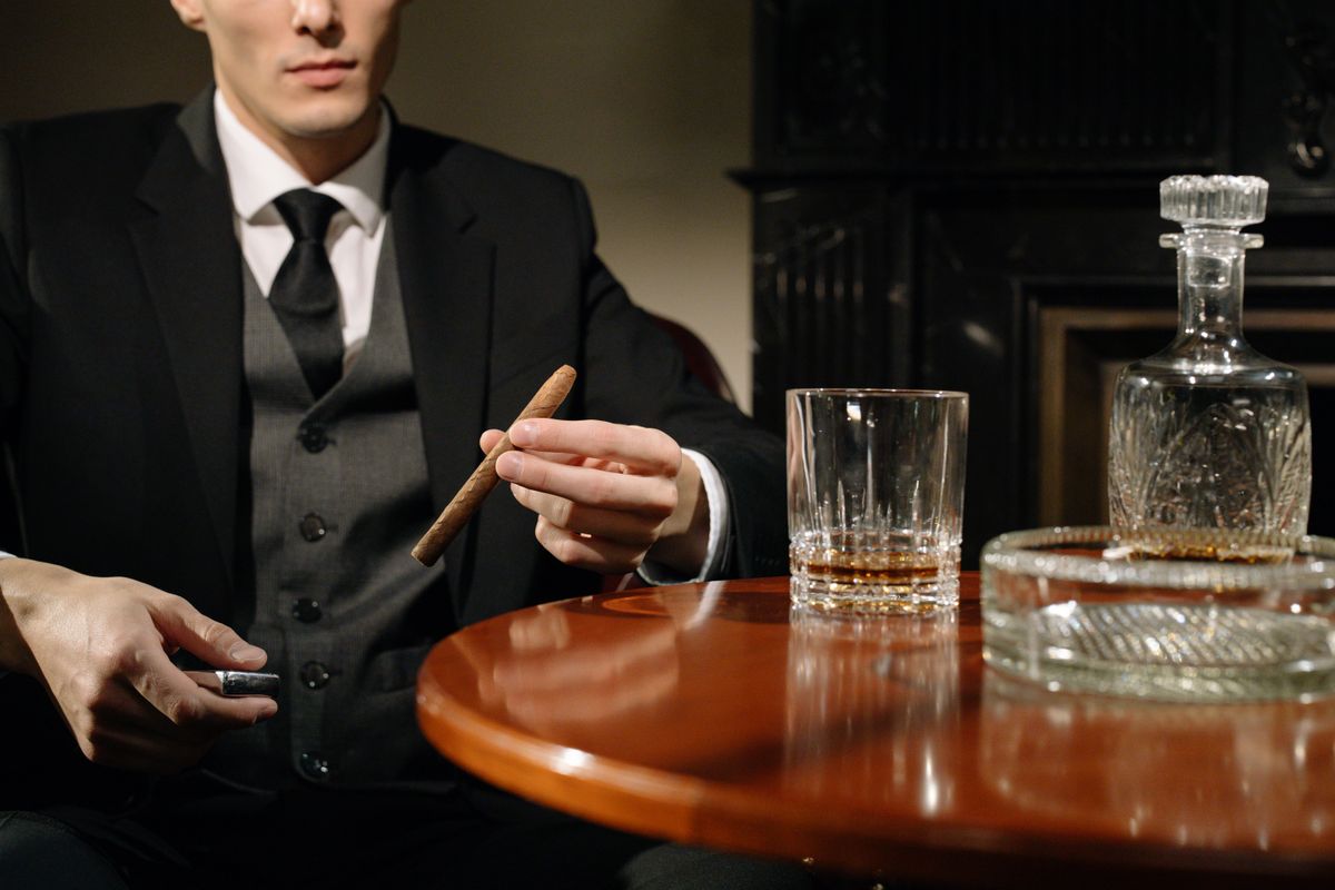 An elegant man in a suit holding a cigar beside a whiskey glass, exuding mystery and class.