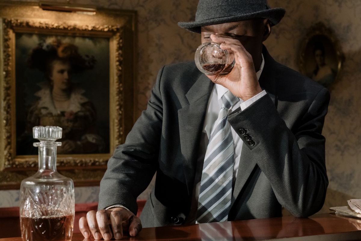 A man in a suit drinks whiskey at a bar, capturing a classic noir atmosphere indoors.