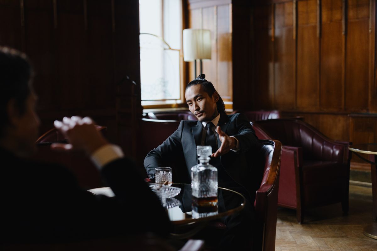 A sophisticated man in a suit sitting indoors with a decanter and drink, exuding style and elegance.
