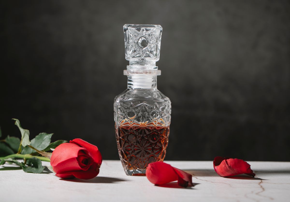 Glass decanter with strong alcoholic drink bourbon placed among rose petals against black background