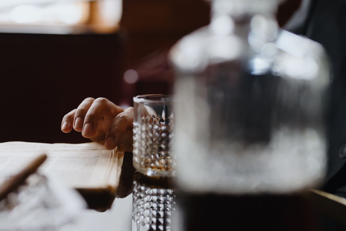 A hand casually rests on a book beside a crystalline whiskey glass in a warm indoor setting.